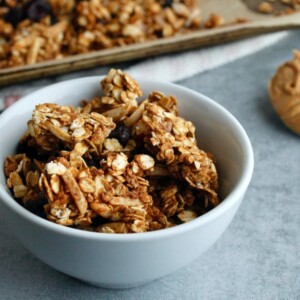 peanut butter granola in a small white bowl; in the background is a white spoon with peanut butter and a tray of baked granola