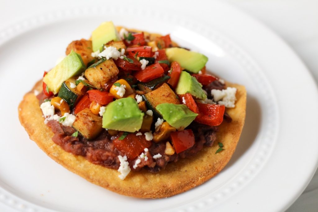 Tostada topped with mashed black beans, a zucchini saute with red peppers and corn, fresh avocado, and a sprinkling of feta