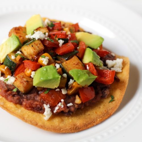 Tostada topped with mashed black beans, a zucchini saute with red peppers and corn, fresh avocado, and a sprinkling of feta