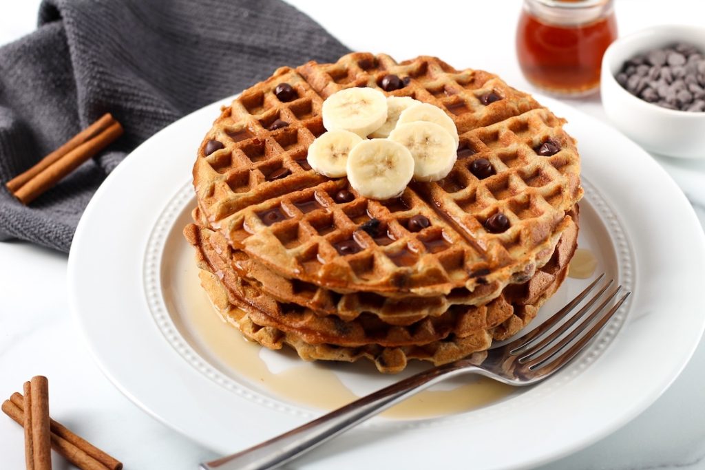 Chocolate chip waffles on a white plate topped with sliced bananas and syrup