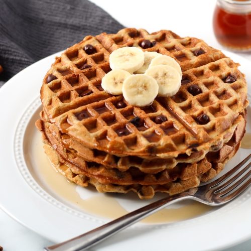 Chocolate chip waffles on a white plate topped with sliced bananas and syrup