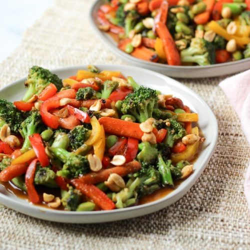 Two plates of teriyaki vegetable stir fry including broccoli, bell peppers, edamame, carrots, and peanuts