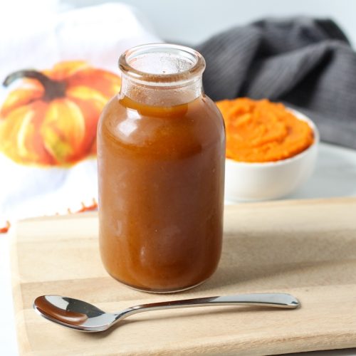 Pumpkin spices simple syrup in a jar on a wooden cutting board. A spoon is in front and canned pumpkin in a bowl and a small pumpkin towel are behind.