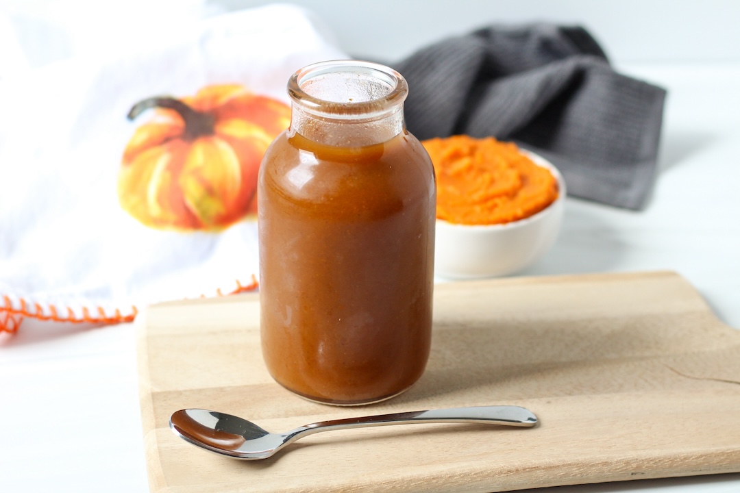Pumpkin spices simple syrup in a jar on a wooden cutting board. A spoon is in front and canned pumpkin in a bowl and a small pumpkin towel are behind.