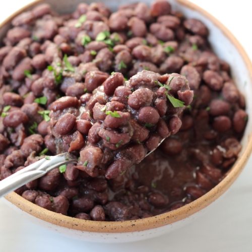 Cuban black beans in a white bowl with a tan rim. Spoonful of beans is in focus while beans in bowl are slightly blurred.