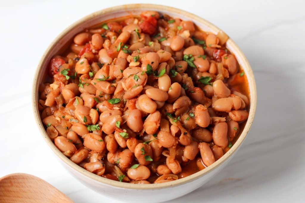 Seasoned pinto beans garnished with cilantro in a white bowl. A wooden mixing spoon is in the bottom left corner.