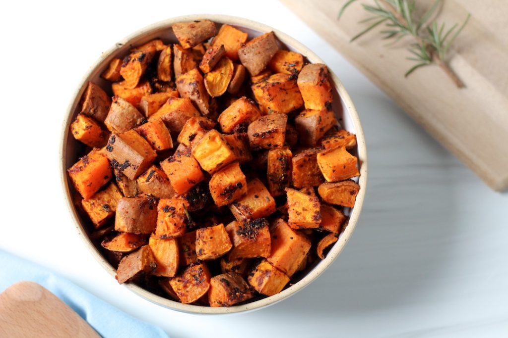 Herb roasted sweet potatoes in a white bowl with a sprig of rosemary nearby.