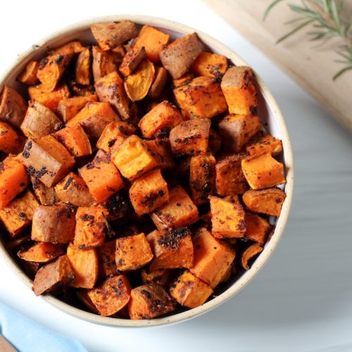 Herb roasted sweet potatoes in a white bowl with a sprig of rosemary nearby.